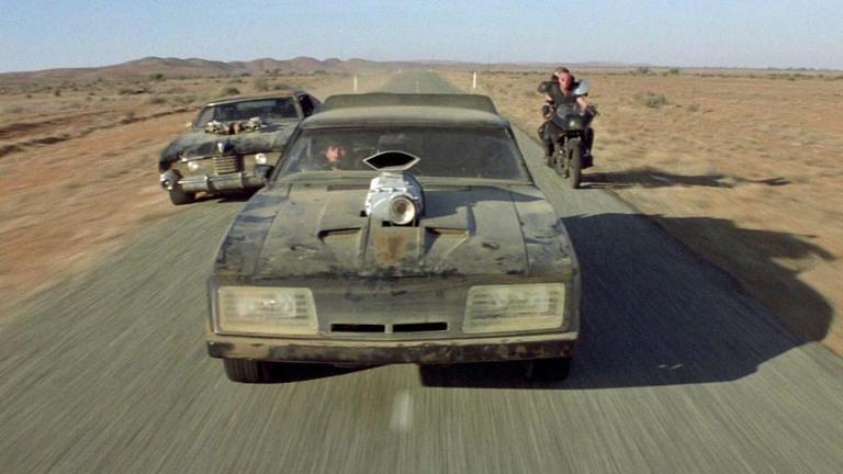 A souped-up car speeding down a desert highway in Mad Max 2, flanked by another car and a motorbike