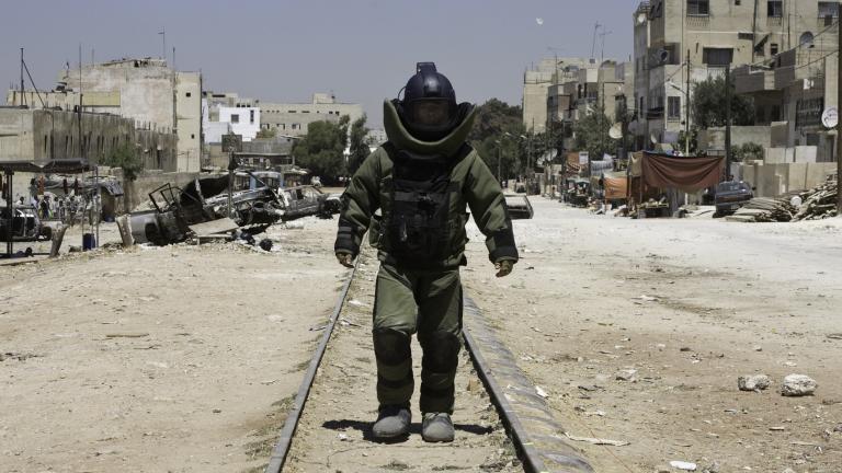 A lonely figure wearing bomb disposal protection walks toward the camera.