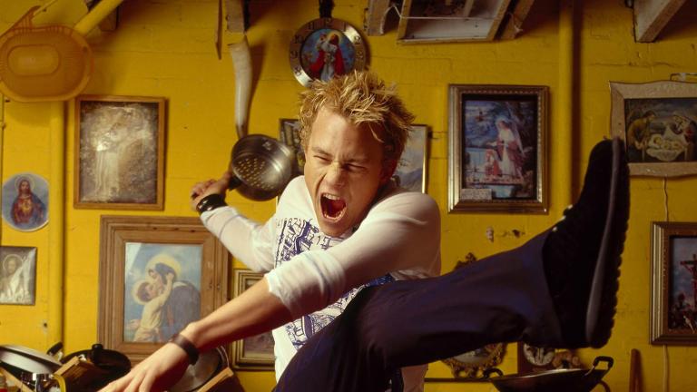 Actor Heath Ledger striking a dramatic pose with his arms out, one leg up in the air and his mouth open wide and eyes shut. He is in a yellow kitchen.