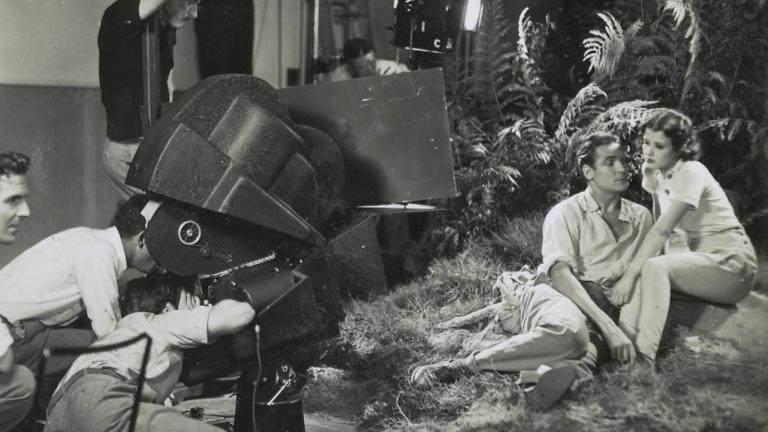 Charles Farrell and Mary Maguire on set sitting together in front of some ferns in front of a crew behind a large film camera Charles Farrell and Mary Maguire on set sitting together in front of some ferns in front of a crew behind a large film camera