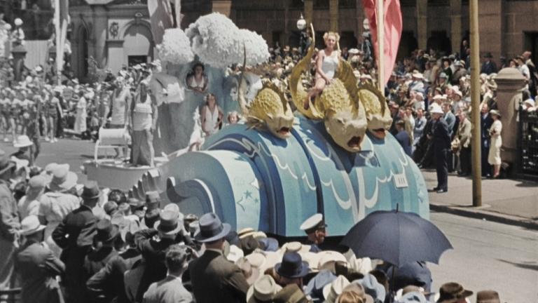 Colourised film still of floats in a parade. Colourised film still of floats in a parade.