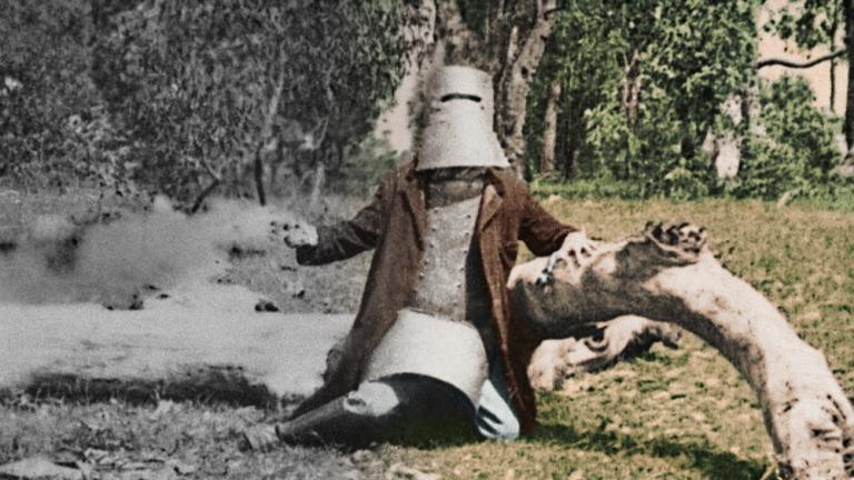Colourised film still of Ned Kelly in armour leaning against a dead tree trunk on the ground. Colourised film still of Ned Kelly in armour leaning against a dead tree trunk on the ground.