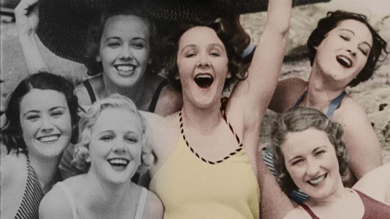 Colourised film still of a group of six women at the beach crowded together under a large hat. Colourised film still of a group of six women at the beach crowded together under a large hat.