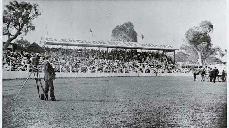 A man standing in an oval with a movie camera. Behind him is a stand full of spectators and on the field is a group of men standing around a horse and carriage. c1900.