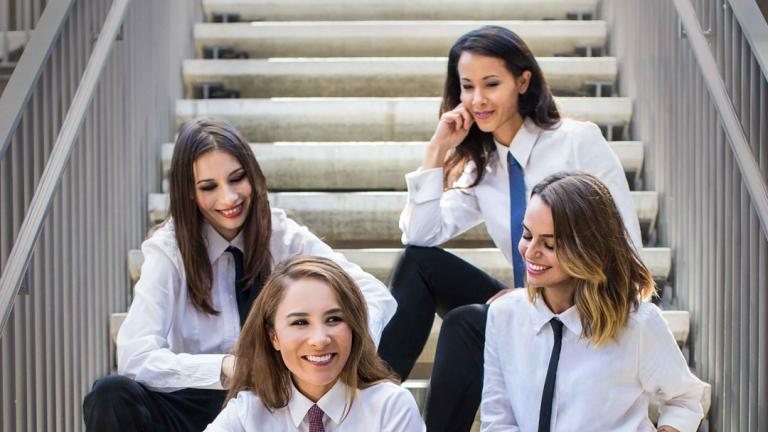Four young women sitting on a staircase. They are all wearing black pants, white button-down shirts and thin neckties.