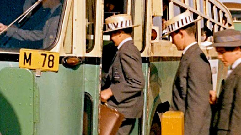 Some schoolboys wearing grey uniforms and hats board a green and yellow school bus.