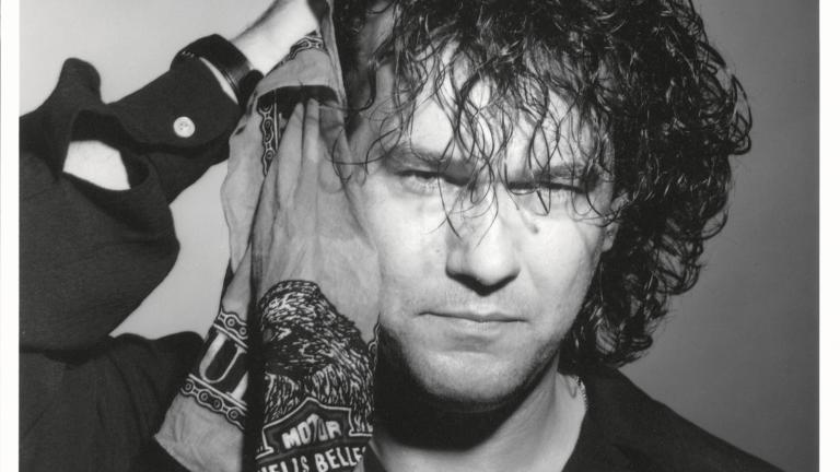 Jimmy Barnes publicity image for Freight Train Heart Head and shoulders shot of Jimmy Barnes wearing a black shirt and looking at camera. He is holding a bandana with a motorcycle club logo on it next to the side of his head.