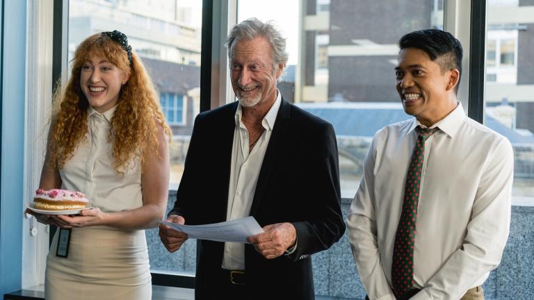 Scene from Halal Gurls featuring Bryan Brown (centre) Three people, a woman and two men, are standing in a row in an office setting smiling. The woman is holding a cake and the man in the centre is reading from a sheet of paper.