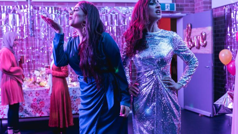 Scene from Halal Gurls Two women dressed in party dresses. One is blowing a kiss to someone left of screen while the other has her hands on hips looking in the other direction. You can see from the background that they are at a party.
