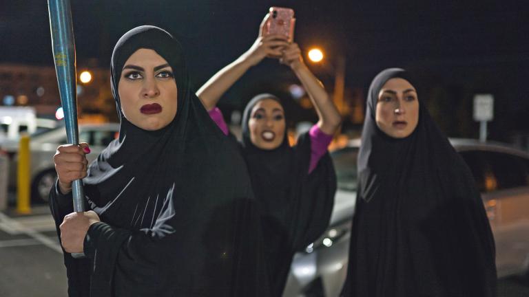 Three women wearing black hijabs in a car park at night time. They are looking angry - one is holding a baseball bat while another is using her mobile phone to video what's happening off camera.