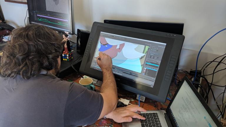 A man pictured from over his shoulder, seated in front of laptop and computer monitor. His screen shows an animated image.