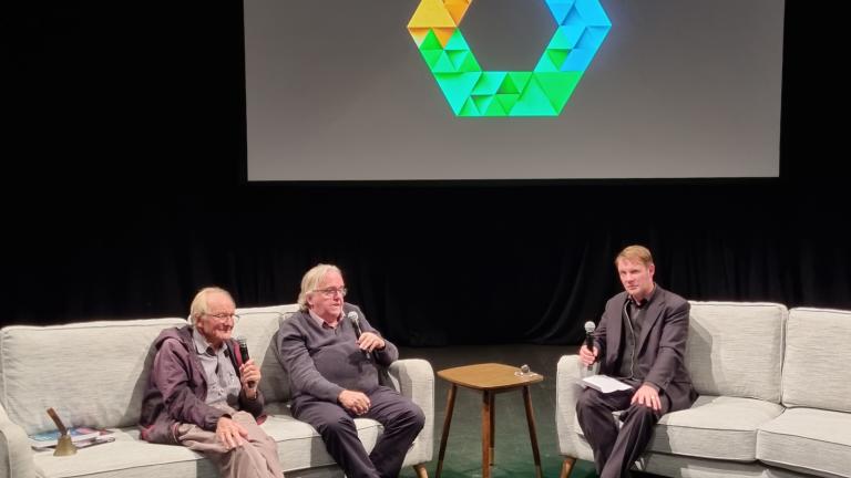 Three men sitting on couches on a stage in a theatre setting. There is a large screen behind them.