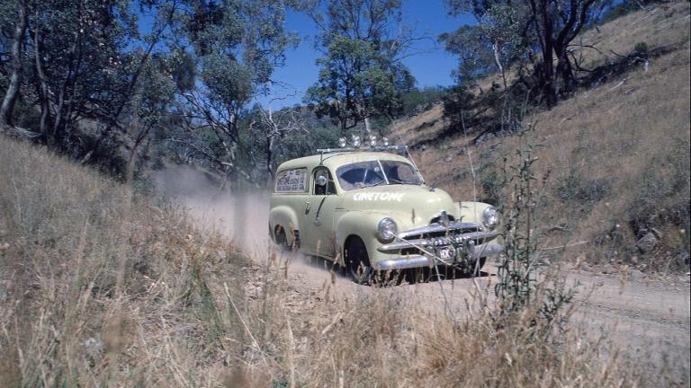 Film still of a cream cvoloured FJ Holden panel van racing along a dusty road.