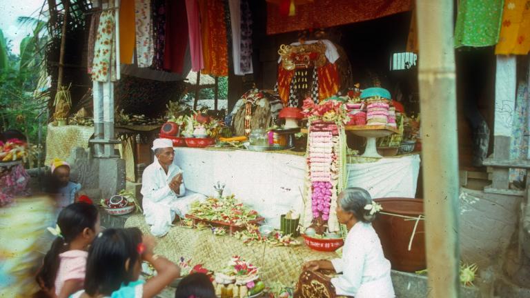 Lempad of Bali - Ceremonial Offerings A Balinese ceremony showing a group of people seated in front of what looks to be a small temple with many bright, colourful offerings at the front.