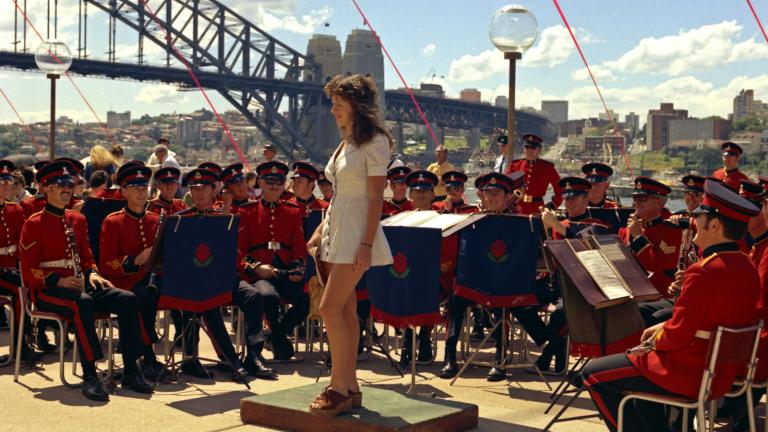 Sydney Opera House Opening - Fifth Facade 3 A woman in a white minidress stands, surrounded by a military band. The Sydney Harbour Bridge is in the background.
