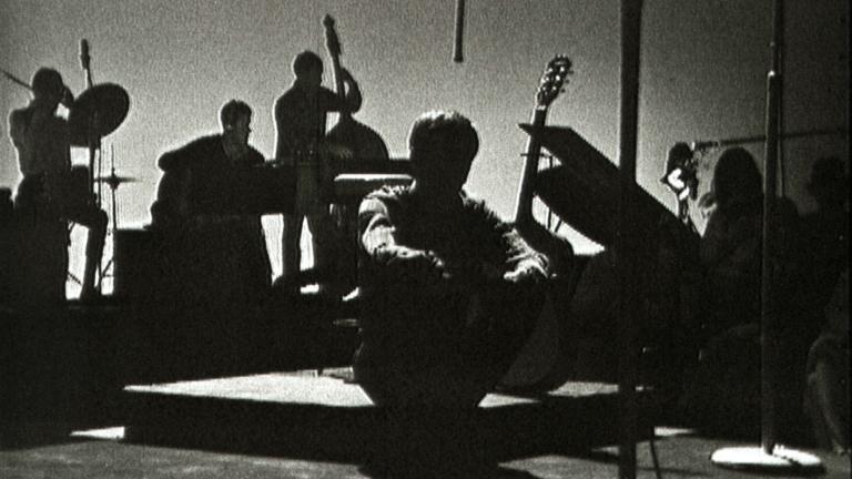Tony Cole on the set of music television show 'Boomeride' sitting in darkness with his backing band behind him.