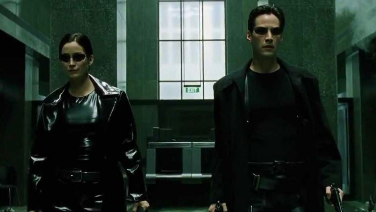 A man and a woman wearing dark sunglasses, black clothes and carrying guns.
