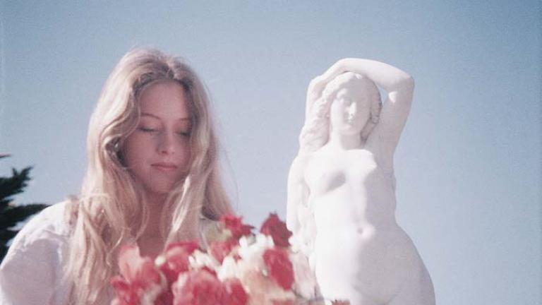 Miranda holds a bouquet of colourful flowers next to a statue of a woman in an outtake from Picnic at Hanging Rock.