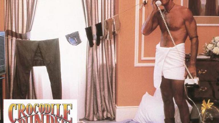 Doing the laundry outback style Lobby card depicting Mick Dundee talking on the phone in his hotel room while he stands wrapped in a towel. He has made a makeshift laundry for his clothes across the window.