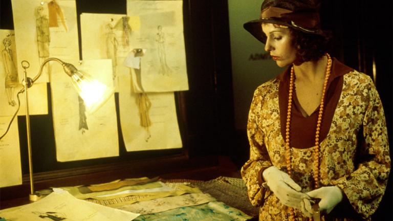 Film still of Helen Morse as Caddie looking at drawings of women's dress designs in a studio.