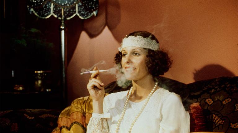 Film still of Helen Morse as Caddie dressed as a flapper, seated and smoking using a long stemmed cigarette holder.