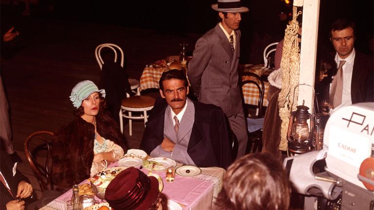 Behind the senes shot of Helen Morse as Caddie and Takis Emmanuel as Peter on the set of Caddie. They sit at a outdoor dining table while the camera crew and extras work around them.
