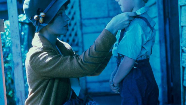 Film still of Helen Morse as Caddie with Sean Hinton as her on-screen son, Terry. Caddie crouching down to straighten Terry's clothes on the verandah of a house.