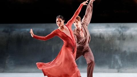 A woman dressed in red and a man dressed in brown dance on a stage against a grey and black backdrop