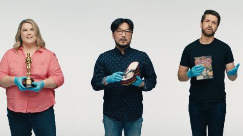 Three people lined up with a white background behind them. The woman on the left holds an Oscar statuette. The middle man holds a platform thong with sequins on it and the man on the right holds a Game CD.