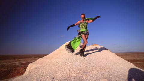 Hugo Weaving in a green dress dancing on a sand dune