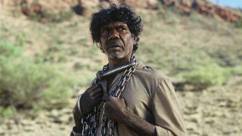 An aboriginal man stands chained in an arid landscape.