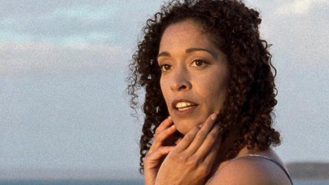 Close up of actor Rachel Maza looking to her left with her hands to her face in a scene from Radiance. The ocean and a cloudy sky can be seen in the background.