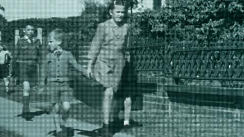 A young woman holding the hand of some school kids walking along a footpath. There are other kids following closely behind them.
