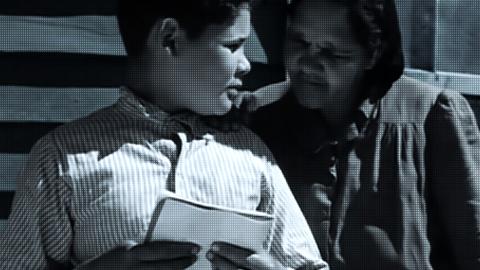 A schoolboy holding a notepad while someone looks over his shoulder at it.