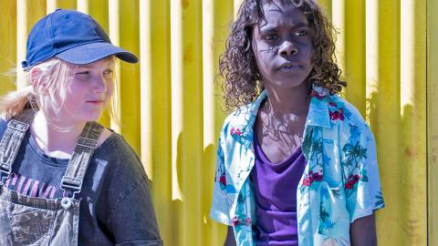 Two kids standing side by side against a yellow corrugated iron fence. One is white girl with straight blond hair in a ponytail, wearing denim overalls and a cap, and the other is a First Nations boy with curly dark hair wearing a purple singlet and blue Hawaiian shirt.