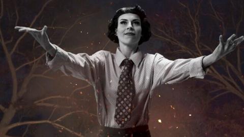 A young woman with short dark hair wearing a business shirt and tie with her arms stretched out wide in front of her. Behind her in the night sky there are tree branches, stars and flickering flames