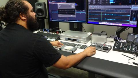 A First Nations man seated at a desk in a studio covered in computers and recording equipment