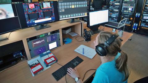 A woman sitting at a desk wearing headphones. She is looking at a number of computer monitors and working on a keyboard. 