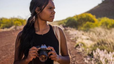 A teenage First Nations girl standing on an outback dirt road holding a camera and looking into the distance.