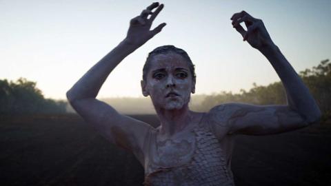 A young woman in body paint doing a dance pose outdoors.