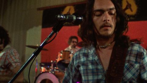 A First Nations man wearing a checked shirt and playing guitar on stage with a rock band in front of an Aboriginal flag.