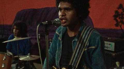 A First Nations man playing guitar on stage with a rock band.