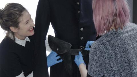 Two curators at the NFSA adjust the belt on a costume on a mannequin. One of the curators is a young woman who is laughing while she works, and both are wearing blue gloves.