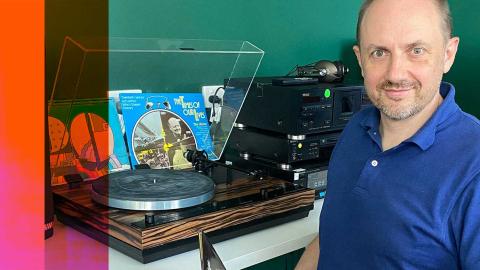 NFSA curator Simon Smith, seated and looking up at the camera. He is sitting at a desk with a turntable and vinyl records on it.