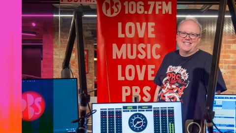 A man stands behind a radio station mixing desk and monitor smiling. He stands in front of a banner that says "106.7FM Love Music Love PBS".