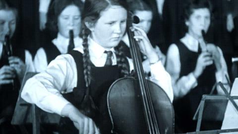 A school girl playing a cello with a group of girls behind her playing recorders.