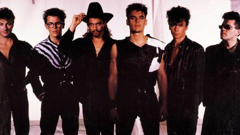 Six members of the band INXS, pictured standing side by side, dressed in black and posing for a publicity image. 