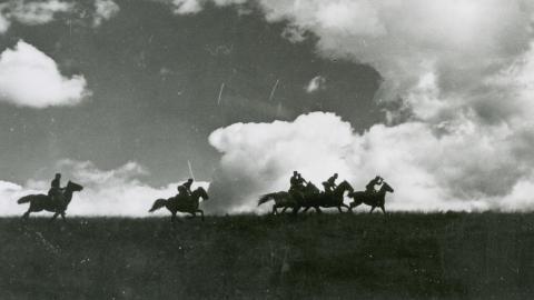Five horses with riders seen in silhouette riding across a flat landscape beneath a cloudy sky