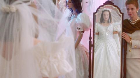A young woman in a wedding dress looking at herself in a full length mirror while an older woman looks on, in a scene from the film Muriel's Wedding. 