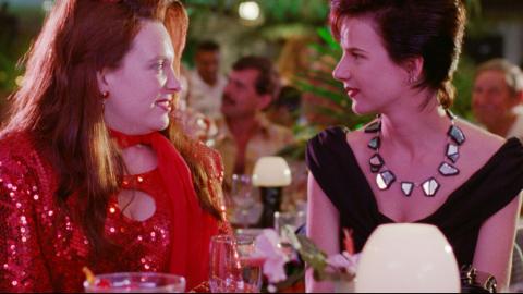 Two women, dressed up for a night out, are sitting at a dining table talking to each other in a scene from Muriel's Wedding.